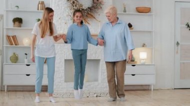 Happy loving intergenerational female family 3 generations having fun weekend together enjoying leisure at home. Young mother and grandmother hold hands granddaughter daughter, girl jumps, does splits