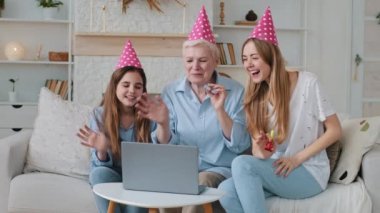Cute child daughter, young adult mother and old grandma looking at laptop screen wearing party hats congratulating to a relative on his birthday remotely due to coronavirus. Family video call concept 