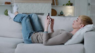 Relaxed young woman lying on sofa in living room holding smartphone using mobile apps. Lazy millennial lady customer surfing social media, shopping, playing game, texting SMS looking at cell at home
