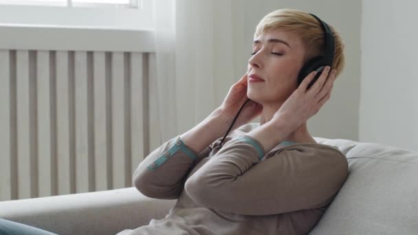 穏やかな若い大人の女性は快適なソファの目にリラックス閉鎖されたウェアのヘッドフォンは、自宅のコンセプトでストレスを感じるリラックス静かな気分を味わう好きなクールな音楽のオーディオブック瞑想をお楽しみください — ストック動画