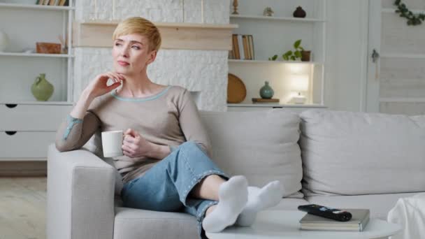 Serene carefree young woman drinking warm tea at home pleasant leisure day. Relaxed calm lady holding cup relaxing sitting on comfortable cozy couch indoors, enjoys good morning feels stress-free — Stok video