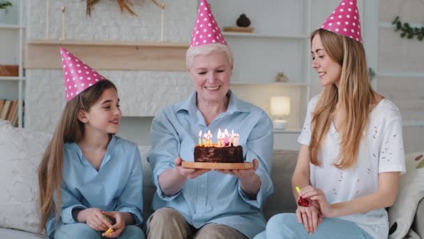 60er Jahre Großmutter mit erwachsener Tochter und süßer lustiger kleiner Enkelin. Alte Oma bekommt von Verwandten Kuchenpusterkerzen, empfindet Dankbarkeit. Weibliche Familie genießen, respektieren und herzliche Beziehung — Stockvideo