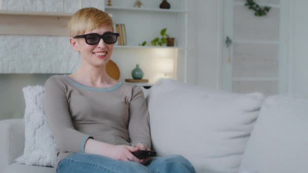 Junge attraktive Frau trägt eine 3D-Brille, hält eine Fernbedienung in der Hand und schaut im Heimkino faszinierende Fernsehfilme, lächelt und genießt das atemberaubende Spektakel auf dem Bildschirm allein drinnen — Stockvideo