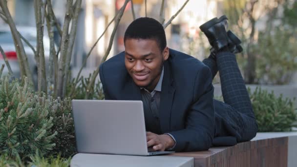Молодой афроамериканский бизнесмен, печатающий на ноутбуке, лежа на животе на открытом воздухе, работая дистанционно, рядом с офисным зданием. Фрилансер смешанной расы Миллениал в костюме, работающий на компьютере на городской улице — стоковое видео