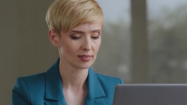 Portrait of beautiful middle-aged Caucasian adult 35s 40s business woman lawyer accountant manager secretary with short hair in green jacket works at laptop online looking at camera smiling friendly