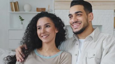 Ethnic spanish couple beautiful brunette woman with curly hair and bearded handsome spaniard man cuddling hugging looking away sitting at home sofa, portrait hispanic satisfied clients smiling teeth