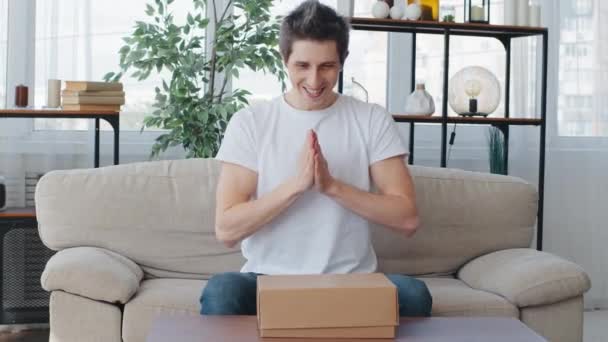 Caucasian man millennial customer buyer sitting on couch rubs palms waiting for gift opens unpacks cardboard box parcel hoping for good product feels disappointed sadness dissatisfaction with delivery — Stok Video