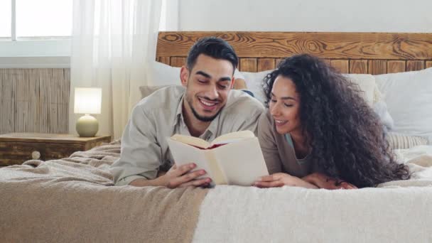 Casal étnico espanhol bela jovem morena com cabelo encaracolado e barbudo bonito espanhol homem deitar juntos na cama ler um livro descansando desfrutar de ler literatura romance em casa no quarto acolhedor — Vídeo de Stock