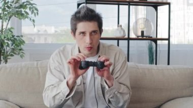 Funny caucasian millennial man adult guy sitting on sofa in home living room comically holding remote controller playing video game console wins resting relaxing lying on couch with hands behind head