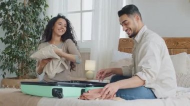 Hispanic latin couple newlyweds spouses curly woman and happy man sitting on bed at home bedroom putting stuff in large suitcase luggage prepare for travel trip abroad vacation guy showing thumbs up
