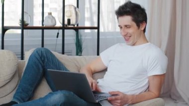 Caucasian millennial cheerful business man adult lies sitting on couch at home looking at laptop screen watching funny humorous video music clip sings laughing loudly sincerely have fun in living room
