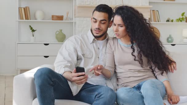 Spanish couple man hispanic husband boyfriend and spaniard woman curly wife girlfriend sitting on couch looking at mobile phone screen read news choose goods online argue quarrel emotionally discuss — Αρχείο Βίντεο