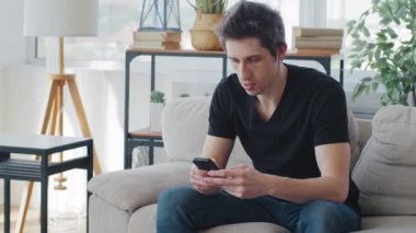 Serious adult Caucasian man wears black T-shirt sitting at home sofa living room looks at mobile phone screen uses smartphone cellphone for chat browsing web orders online delivery receives message