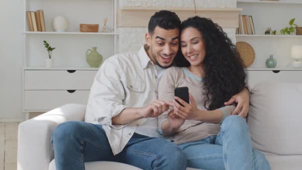 Famiglia ispanica coppia coniugi sposi spagnolo uomo e giovane donna abbracciare abbraccio uso telefono guardando lo schermo dello smartphone ridendo sinceramente sorridente con divertente app mobile seduto sul divano — Video Stock