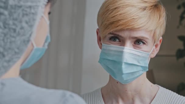 Close-up female masked face, portrait caucasian sick woman patient wears medical protective mask speaks to unrecognizable lady doctor nurse practitioner talks about disease symptoms consult in clinic — Stock Video