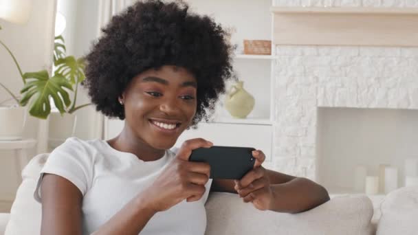 Happy relaxed millennial mixed race girl holding smartphone looking at cellphone laughing enjoy using mobile apps for shopping having fun playing games chatting in social media sit on sofa — Stok Video