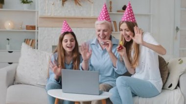 Smiling young adult mother, cute adorable child granddaughter and older mature grandmother in funny party hats congratulating relative birthday communicate video calling chatting by webcam app