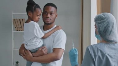 Young serious man afro american father holds in hands little African daughter child preschool girl puts palm in front of him refuses prohibits vaccination injection with vaccine to woman doctor nurse
