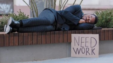 Depressed unhappy desperate mixed race office worker, black young adult businessman lying outdoors awaiting offer. African American student millennial cant get employment, showing poster need work