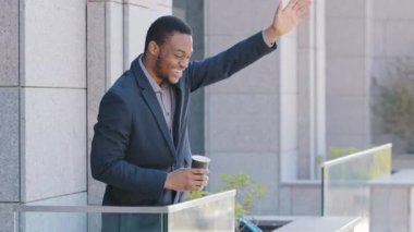 Smiling young confident African American businessman standing on balcony of office enjoying rest during break. Mixed race millennial guy wearing suit drinking coffee, waving hand greeting colleague