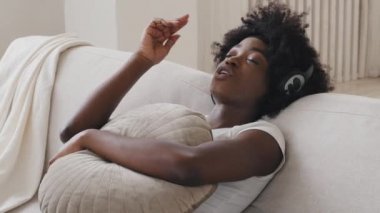 Calm happy young black race woman in headphones sitting on comfortable sofa with eyes closed listening favorite music modern rhythmic pleasant composition enjoy peaceful mood wearing earphones at home