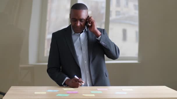 Starší středního věku africké 50s obchodní muž šéf vůdce ve formálním obleku mluvit na mobilním telefonu s klienty poradenství s mobilním telefonem smartphone stojící v kanceláři psaní lepkavé poznámky na samolepky
