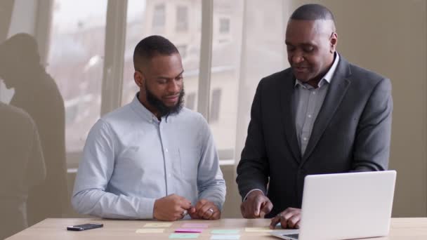Два африканских американских бизнесмена афро-взрослый лидер босс и черный этнический менеджер работают вместе стоя за столом с ноутбуком в офисе мозговой штурм обсуждая идеи писать на липких нотах — стоковое видео