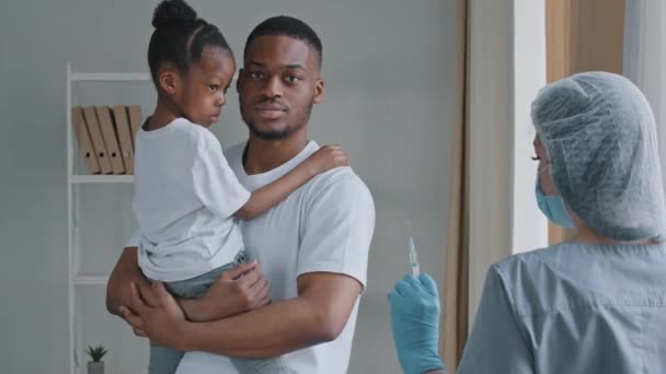 Giovane uomo serio afro-americano padre tiene in mano la piccola figlia africana bambina prescolastica mette palmo davanti a lui rifiuta proibisce la vaccinazione iniezione con vaccino alla donna medico infermiere — Video Stock