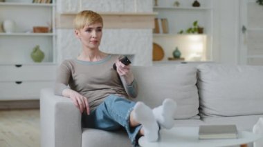 Woman switching channels choose comedy film for funny pastime, relaxing on comfortable cozy sofa indoors alone, watching entertaining programs reality show series on TV, spending leisure time inactive