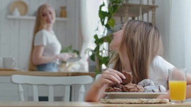 Cute teenage girl child daughter sitting at table in kitchen at home breakfast in morning eating delicious sweet homemade chocolate cookies with juice talking laughing with adult mother washing dishes