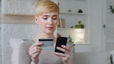 Woman customer holding credit card and smartphone sitting on couch at home. Female shopper using instant easy mobile payments making purchase in online store receiving cashback. E-banking app service