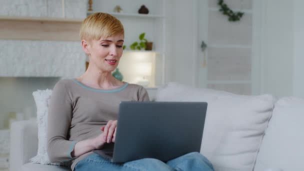 Mujer joven bastante feliz acostada en un sofá acogedor con computadora portátil en vueltas, trabajando lejos de casa, escribiendo correo electrónico, disfrutando de las compras en línea o comunicándose en las redes sociales, sintiéndose relajada el fin de semana — Vídeos de Stock