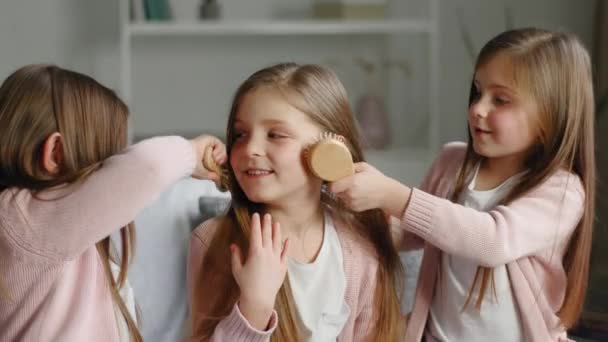 Tres graciosas niñas lindas hermanas mejores amigos trillizos gemelos mismos niños sentados en el sofá de casa dos niños peinando el pelo largo y suave de una colegiala usando peines cepillado hacer peinado peinado coiffure — Vídeo de stock