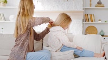 Genç aile şefkati, beyaz anne, bebek bakıcısı anne, kanepede kızıyla oturuyor, anaokulundaki küçük kız, saçını tarıyor, saçını kestiriyor, her sabah tarama ve güzellik prosedürleri uyguluyor.
