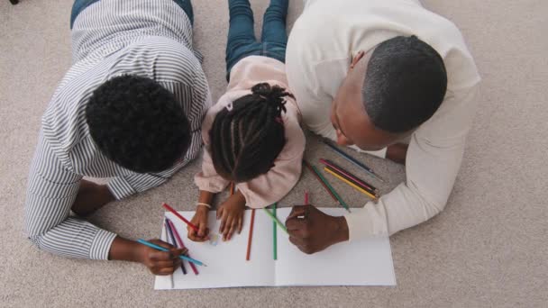 Вид сверху неузнаваемая африканская американская семья афро черные родители с этнической дочерью школьница талантливый творческий ребенок лежат на домашнем этаже рисовать картину вместе с помощью цветных карандашей рисования хобби — стоковое видео