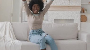 Mutlu, rahat, Afro-Amerikan, genç, yetişkin siyahi kadın evinde rahat bir kanepede uzanıp uzaktan kumandayı tutarak spor kanalı TV programını izlerken heyecanla kazanıyor. Futbol maçı fan konsepti