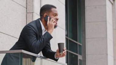 Ciddi özgüvenli sakallı Afrikalı Amerikalı bin yıllık iş adamı cep telefonuyla konuşuyor. Ofis balkonunda duruyor. Siyahi erkek yönetici tatilde cep telefonuyla konuşuyor.