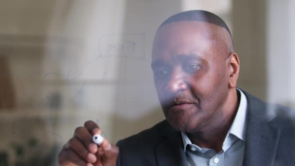 Pohled za sklem africký americký dospělý muž afro černošky obchodní šéf vědec učitel píše na transparentní palubě v kanceláři dělá poznámky s marker řeší problém přemýšlí o strategii — Stock video