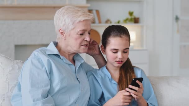 小さなかわいい孫娘が手にスマートフォンを保持している古い祖母は、モバイルアプリケーションを使用してゲームをプレイし、写真を示しています。高齢者ベビーシッターは、携帯電話上の教育ビデオと女の子を助ける — ストック動画