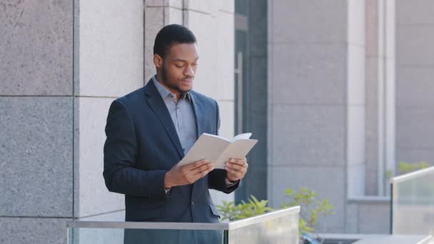 Grave uomo di colore sicuro che indossa vestito che tiene pianificatore giorno, lettura libro di carta o taccuino d'affari rilassante sul tempo libero. Uomo d'affari africano in piedi sul balcone di ufficio godendo di riposo durante la pausa — Video Stock