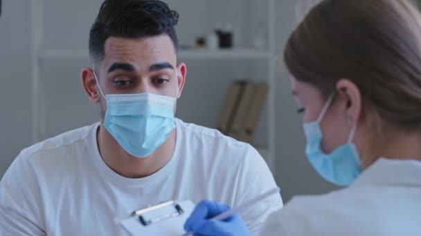 Arabic man hispanic guy patient sick male wears medical protective mask on face talks about symptoms of health problems to unrecognizable doctor woman nurse writing down notes, consultation in clinic — Stock Video