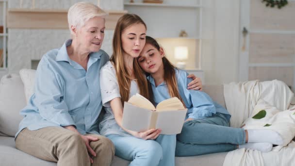 Glücklich liebende Familie drei Generationen verbringen das Wochenende zusammen auf der Couch im gemütlichen Wohnzimmer sitzen. Junge Mutter liest Lieblingsbuch für Tochter und Großmutter. Konzept familiärer Traditionen — Stockvideo