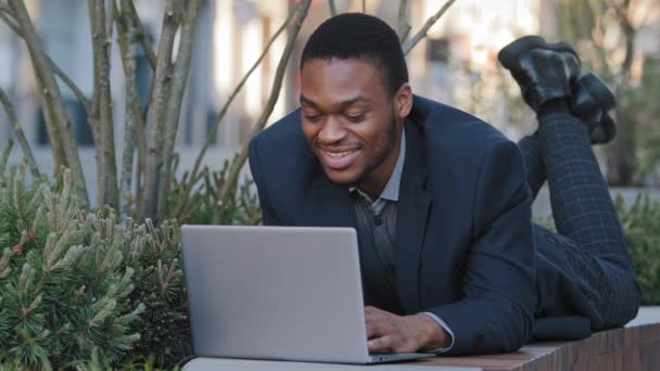 Lachend zelfverzekerd tevreden jonge volwassen zakenman of zakelijke werknemer liggend maakt gebruik van laptop, communiceert in sociale netwerken voorbereiding van spraak. Gemengde race zwart millennial student studeren outdoor — Stockvideo