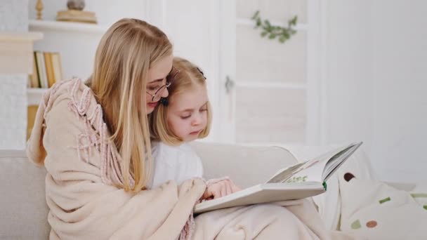 Joven cariñosa madre caucásica lleva gafas cubiertas manta sentado en el sofá de casa con la niña pequeña niña preescolar sosteniendo libro de fotos muestra imágenes cuenta historias comparten recuerdos — Vídeo de stock