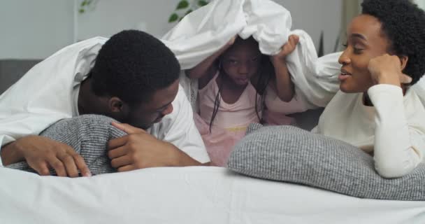 Young afro american parents black mother and father lying on bed sofa at home talking chatting conversation little ethnic african daughter child girl hiding under blanket man looking at mobile phone — ストック動画