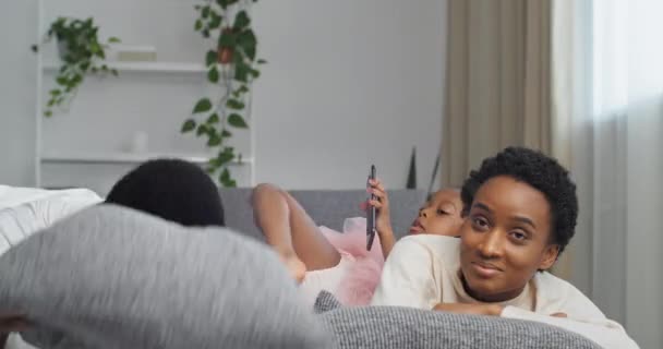 Afro american family black ethnic african parents lying on couch resting man father hiding under pillow little daughter girl child looking at a mobile phone playing game on smartphone using cellphone — Stock Video