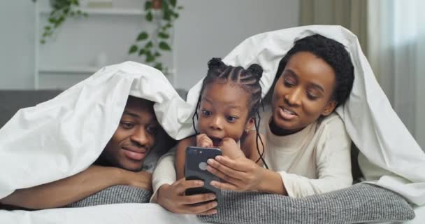 Afro-Amerikan komik ailesi beyaz battaniyenin altında kanepede uzanmış fotoğraf maskelerinin online uygulaması için cep telefonu kullanıyor ve gülümsüyor. — Stok video