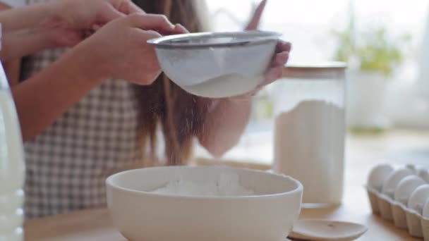 Närbild två par kvinnliga händer oigenkännlig kaukasiska mor och dotter hjälpare tillsammans hålla järn sikt sålla mjöl i skålen förbereda deg med ägg och mjölk för kaka paj bröd i köket — Stockvideo