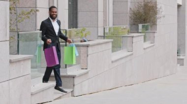 Alınan güzel şeylerden mutlu, Afrika kökenli sakallı iş adamı piyasaya çıkıyor. Satın almaktan memnun gülümseyen, alışveriş sırasında renkli alışveriş torbaları taşıyan bin yıllık melez insan.