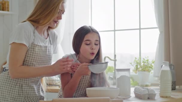 Madre adulta con el pelo largo enseña a cocinar pequeña hija adolescente tamizar harina juntos a través de criba de hierro niño soplando en el aire riendo preparaciones divertidas cocinar en la cocina casera antes del desayuno — Vídeos de Stock